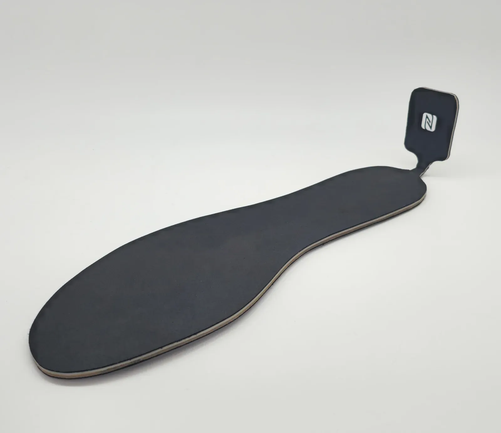Black fabric-topped insole lying on a white surface, with a small NFC tab on a narrow neck at the heel